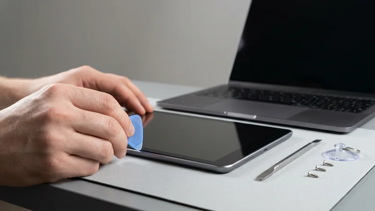 Hands ease a thin plastic opening pick along the seam of a tablet lying beside an open laptop on a repair bench, both screens dark, with a spudger and loose screws laid out alongside.