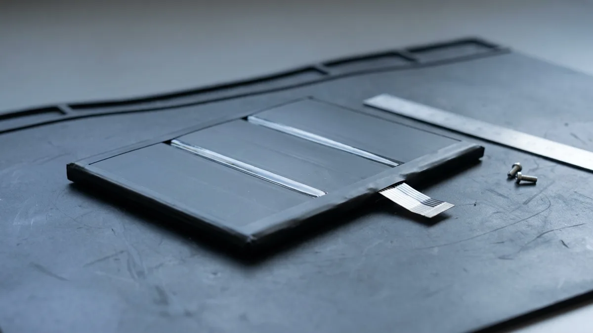 A single slim black laptop battery pack lies flat on a scuffed dark bench mat, three long flat cells showing through its matte wrap and a short pale ribbon lead folded across one corner, with a plain metal rule and two small screws beside it.