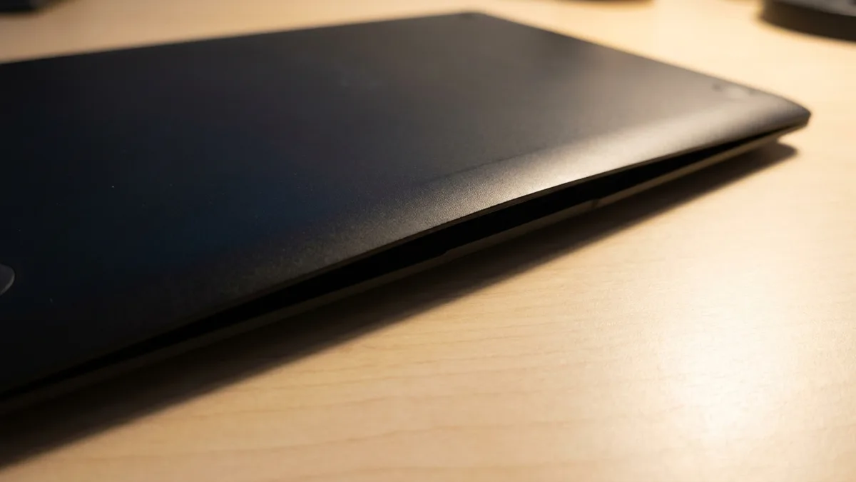 The closed underside of a thin dark laptop on a pale wooden desk, its bottom cover no longer sitting flush: the panel has bowed upward across the middle and a dark tapering gap runs along the seam where the cover meets the frame.