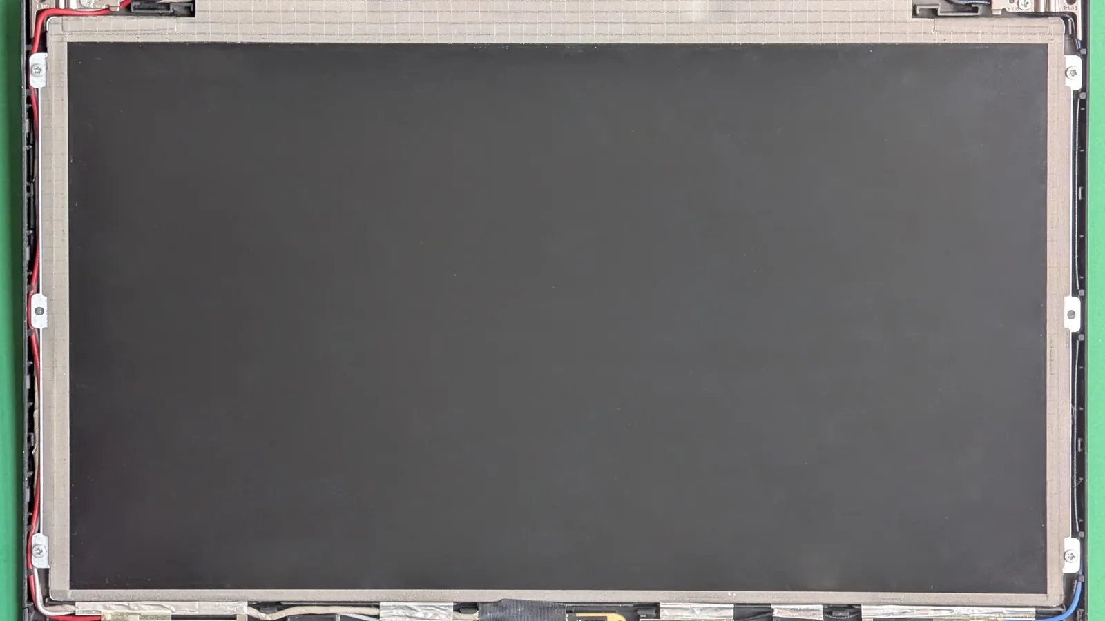 A bare laptop LCD panel lifted from its lid and seen from the front, its dark blank screen filling a silver metal frame; small screw brackets and clips grip the edges, red and black wires run down the left side, and strips of metallic tape and cabling cross the bottom, all on a green mat