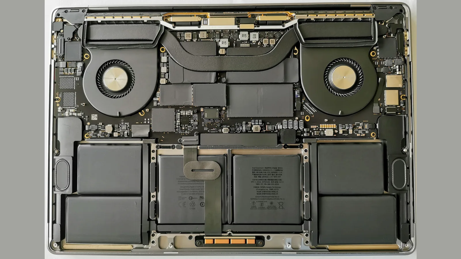 The inside of a 16-inch MacBook Pro with the bottom case removed, seen from directly above on a grey background: a round cooling fan in each upper corner, a black heat pipe curving across the top between them, the logic board studded with small chips and printed codes, six flat battery cells filling the lower half, and a speaker module at each outer edge