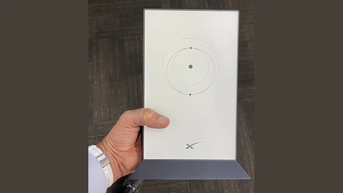 A hand holding a white rectangular SpaceX Starlink user terminal on its grey stand