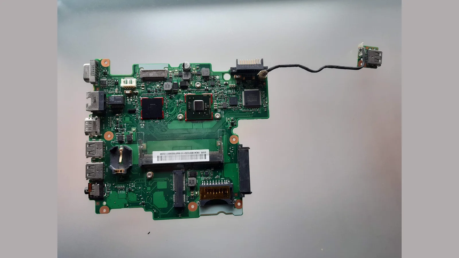 Papan induk komputer riba berwarna hijau yang dikeluarkan dari kotaknya, terletak di atas permukaan kelabu: port di sepanjang tepi kirinya, slot memori berlabel putih, cip-cip, dan papan USB kecil pada kabel di sebelah kanan atas.