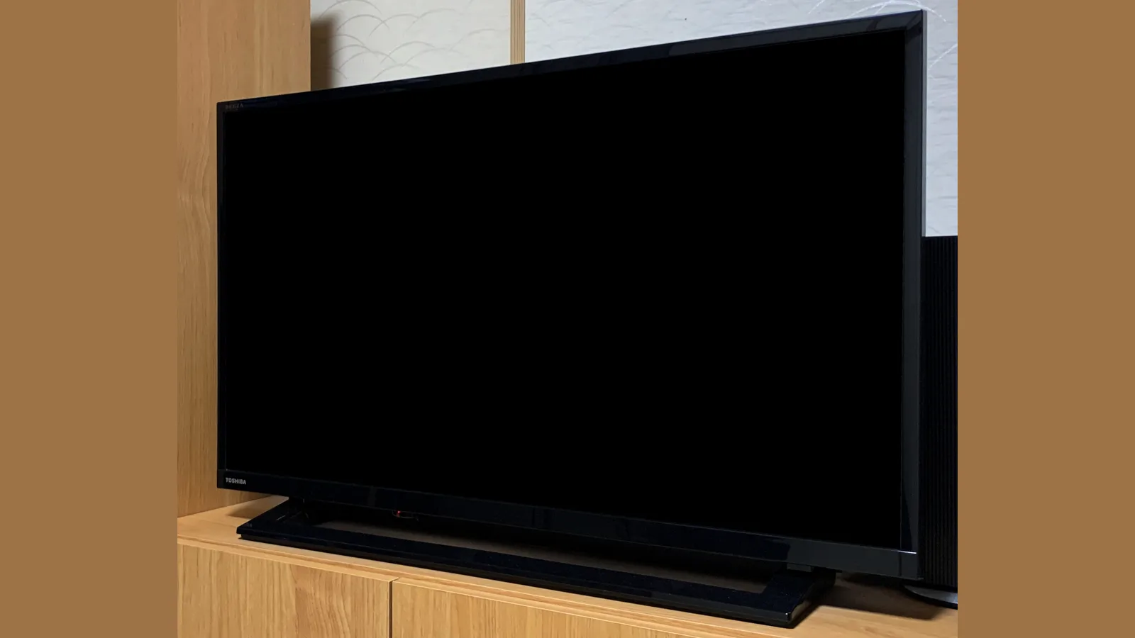 Sebuah TV skrin rata Toshiba berwarna hitam di atas kabinet kayu cerah, skrinnya gelap, dengan nama TOSHIBA di sebelah kiri bawah bingkainya yang nipis.