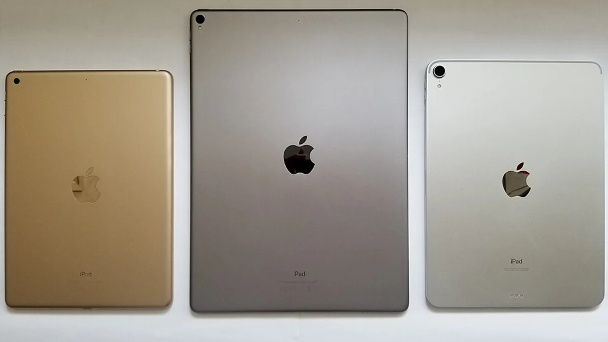 Three iPads of different sizes lying face-down side by side on a plain white surface, in gold, space grey and silver, each with the Apple logo and the word iPad on the back.