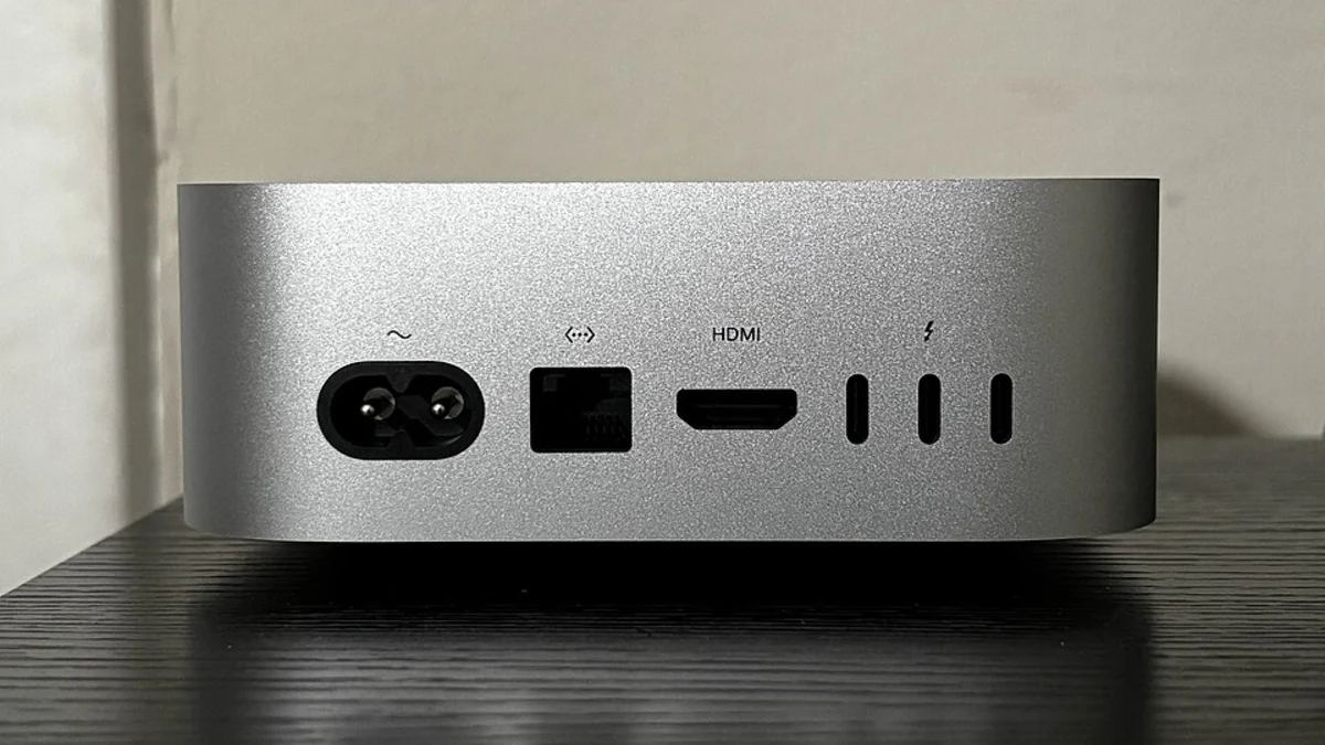The back panel of a silver Mac mini standing on a dark wooden surface, showing the power inlet, an Ethernet port, an HDMI port and three USB-C ports in a row.