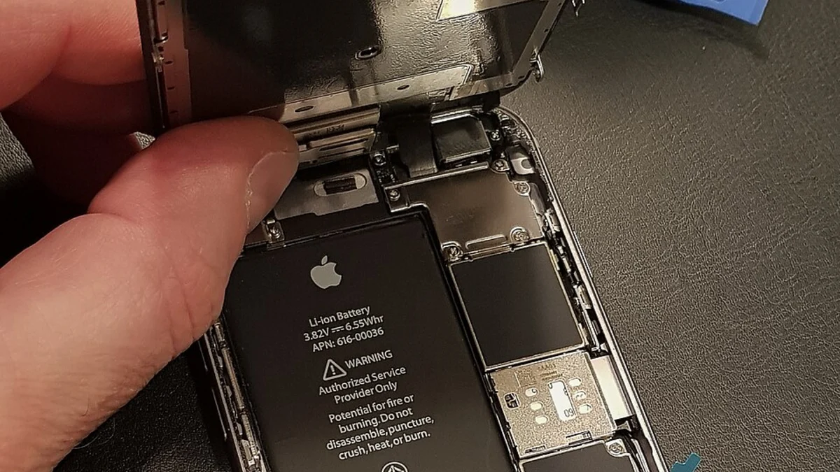 A hand lifting the display assembly away from an opened iPhone, revealing the genuine Apple lithium-ion battery in the chassis with its printed warning label, and a blue plastic opening tool at the top of the frame.