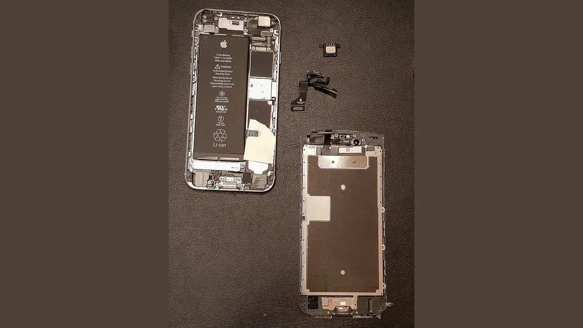 An opened iPhone chassis with its genuine Apple battery still fitted, laid on a dark mat beside its removed display assembly, with the earpiece speaker and the front-camera flex cable set out between them.