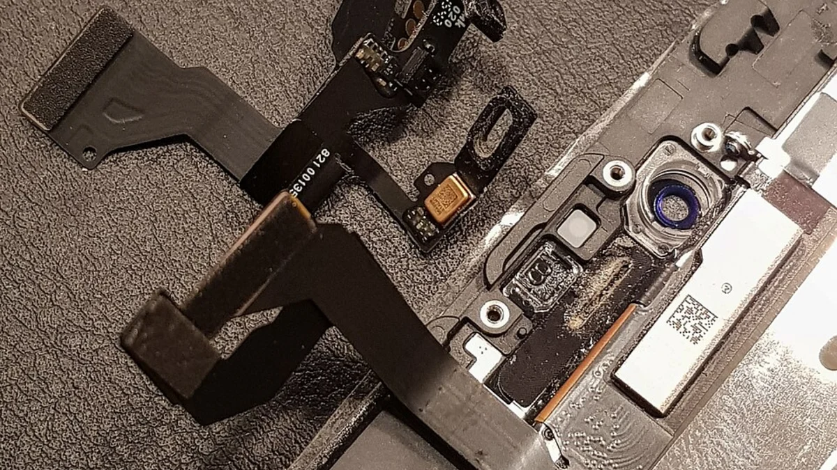 The front-camera and proximity-sensor flex cable lifted clear of an opened iPhone and laid on a dark mat, its ribbon folded back beside the metal display frame it came from.