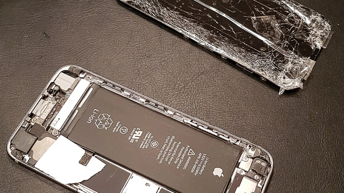An opened iPhone chassis with its genuine Apple battery in place on a dark mat, beside the shattered display it has just had removed, with repair tools laid out at the top of the frame.