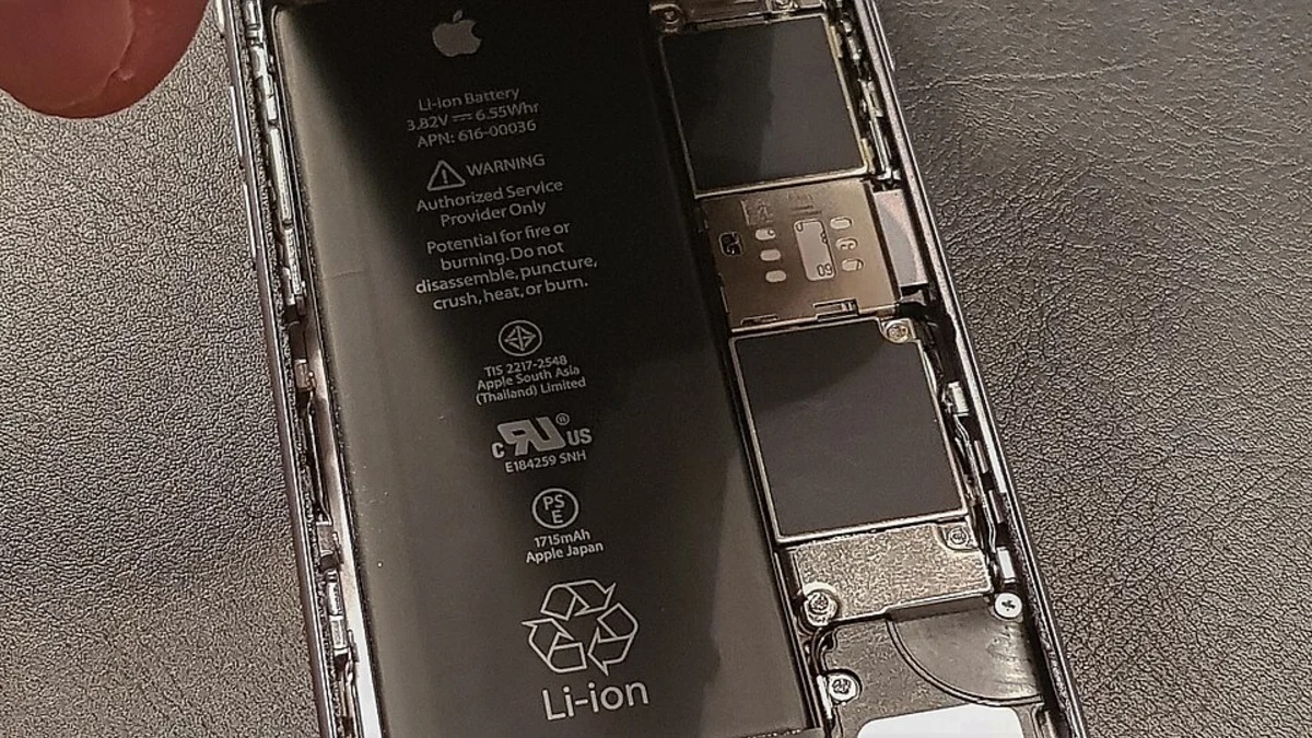 A close view of the genuine Apple lithium-ion battery inside an opened iPhone, its printed label giving the voltage, the watt-hour rating and the part number, with a fingertip at the edge of the frame.