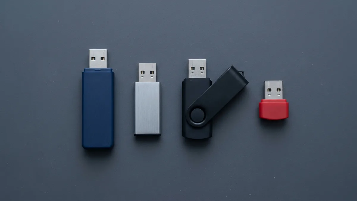 Seen from above on an otherwise empty dark grey surface: four unmarked USB flash drives standing side by side with their metal plugs uncapped and facing up the frame — a long dark blue one, a slim brushed-silver one, a black one with its swivelling cover rotated open, and a short red stub.