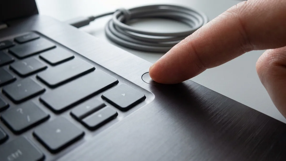 A fingertip pressing the round power button beside the keyboard on a laptop's brushed-metal palm rest, with a coiled cable lying unplugged on the desk behind