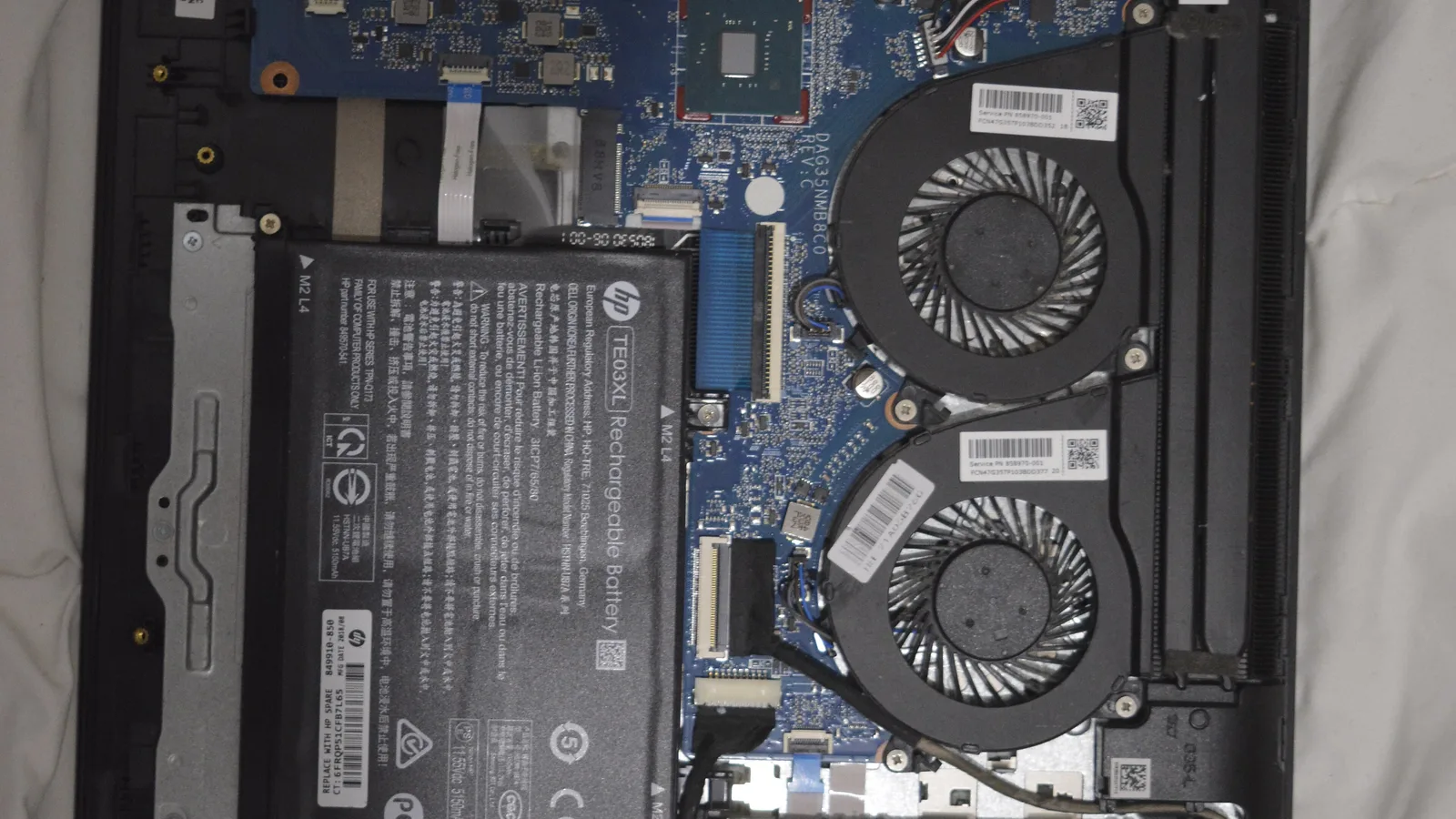 The inside of an opened laptop with the bottom cover removed, showing a black HP battery pack with printed text on the left, a blue circuit board across the middle, and two round cooling fans set in a dark housing on the right, on a pale cloth background.